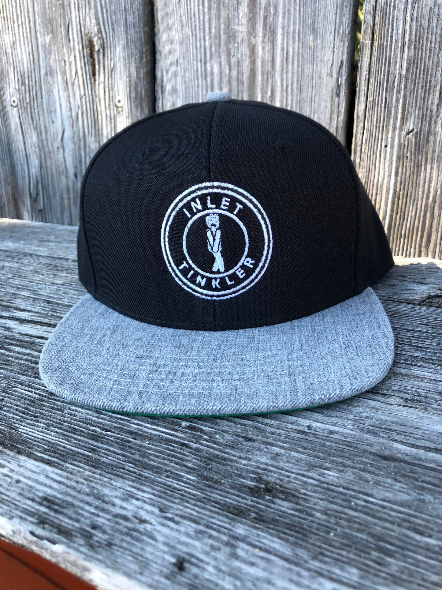 Black and grey flat bill snapback hat with embroidered Inlet Tinkler logo on the front photo taken at marshwalk