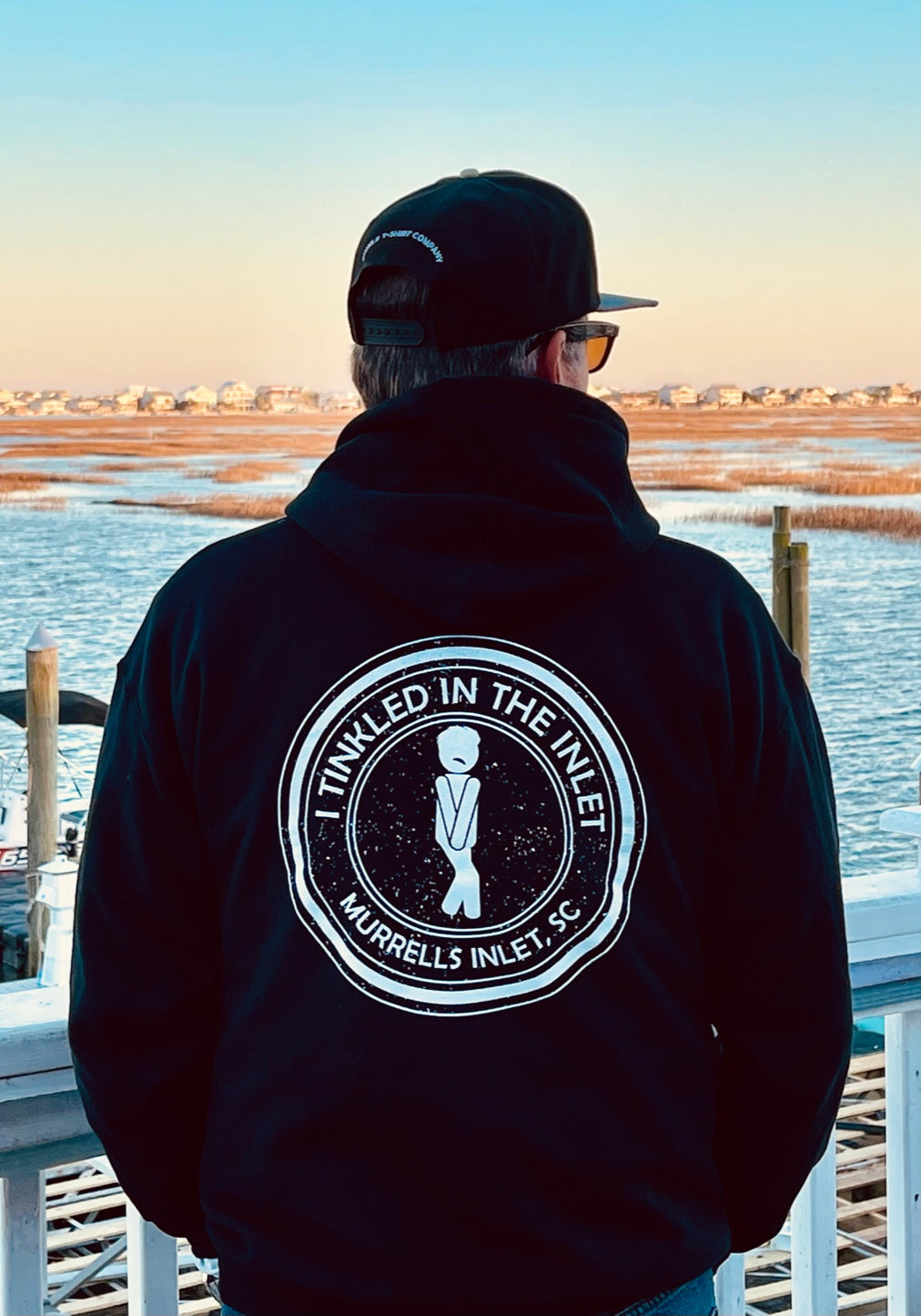 man standing at marshwalk at murrells inlet wearing a black "I Tinkled in the Inlet Hoodie" facing the water.