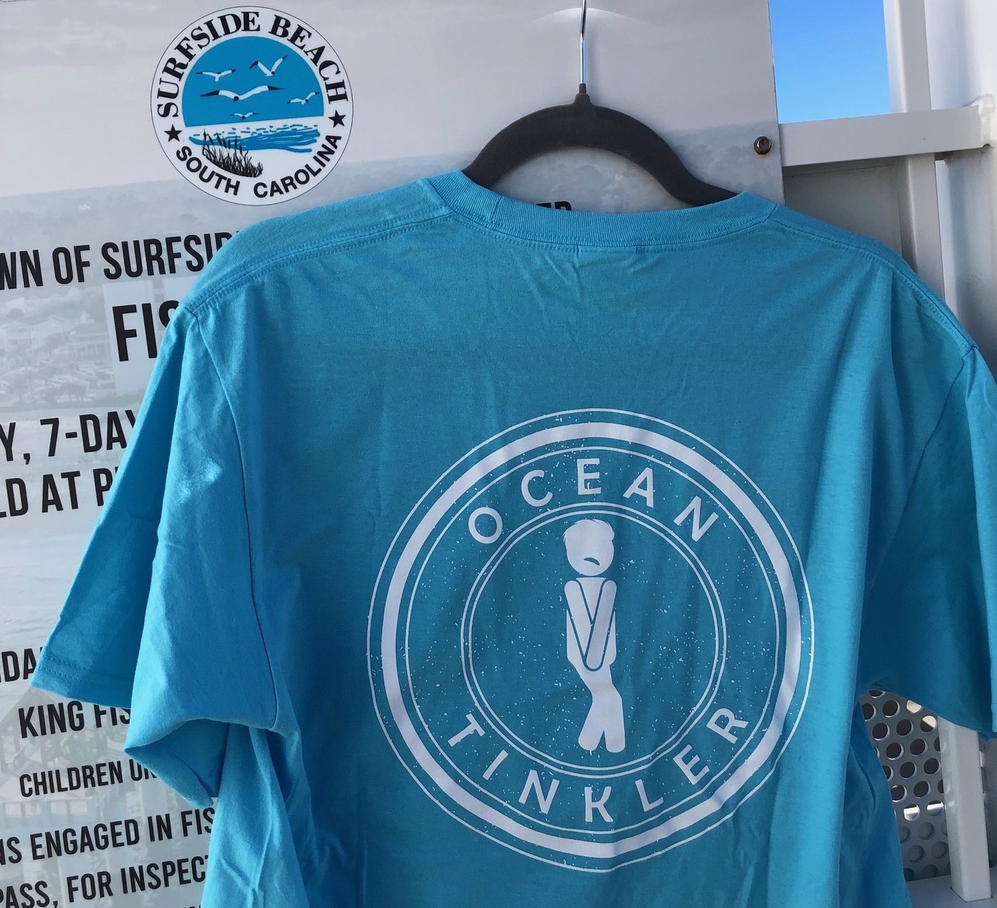Back of t-shirt with white Ocean Tinkler graphic on aqua blue fabric hanging in surfside beach pier
