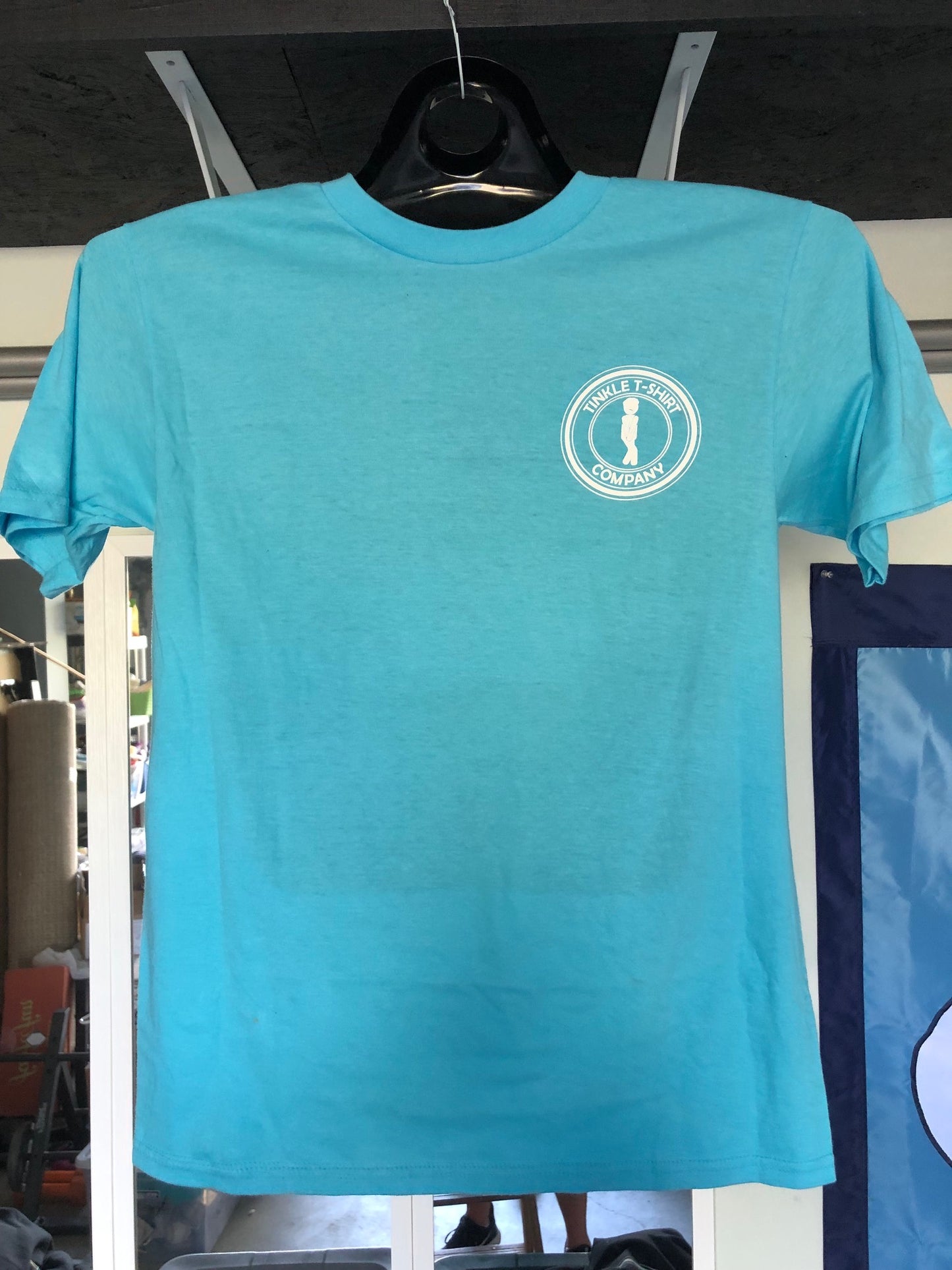 Front of aqua blue t-shirt with Tinkle T-Shirt Company logo on hanger